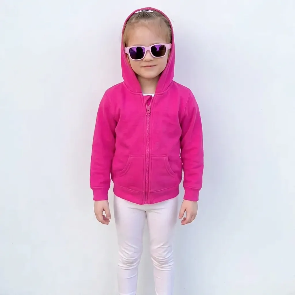 Girl in a pink hooded jacket, light trousers and sunglasses against a white background.